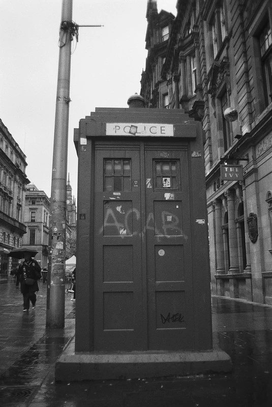 Schwarz-Weiß-Fotografie einer britischen Polizeikabine (Police Box) auf einem städtischen Gehweg. Die Tür der Kabine ist mit dem Graffiti 'ACAB' beschriftet. Im Hintergrund sind historische Gebäude und Passanten auf einer regennassen Straße zu sehen.