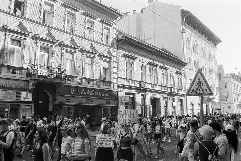 Schwarz-Weiß-Fotografie der Budapest Pride 2023: Eine Menschenmenge läuft durch eine Straße zwischen historischen Gebäuden. Im Vordergrund hält eine Person ein Schild mit der Aufschrift 'Protect Trans Kids' hoch.