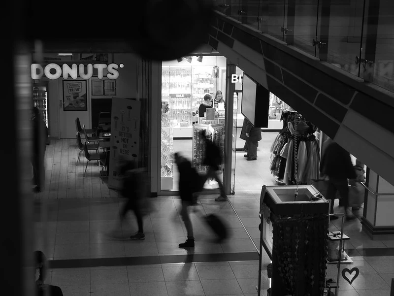 Schwarz-Weiß-Fotografie eines Bahnhofs oder Einkaufszentrums: Im Hintergrund ist ein Donut-Laden mit beleuchtetem Schild zu sehen. Menschen sind in Bewegung, einige leicht verschwommen, was eine dynamische Atmosphäre erzeugt. Im Vordergrund sind eine Rolltreppe und ein Mülleimer zu erkennen.