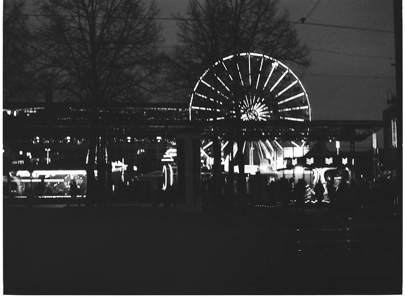 Schwarz-Weiß-Nachtaufnahme eines Rummelplatzes: Im Zentrum leuchtet ein großes Riesenrad, umgeben von weiteren Fahrgeschäften und Ständen. Im Vordergrund sind Bäume und Stromleitungen zu erkennen, die eine stimmungsvolle Atmosphäre erzeugen.