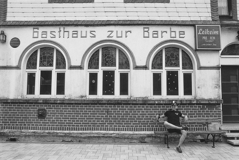 Schwarz-Weiß-Fotografie eines traditionellen Gasthauses namens 'Gasthaus zur Barbe': Im Vordergrund sitzt eine Person mit Sonnenbrille auf einer Bank und lächelt direkt in die Kamera.