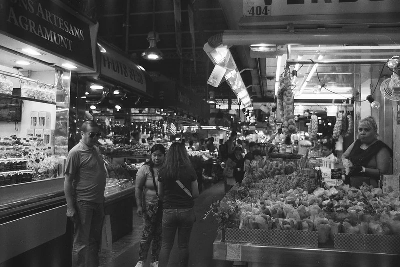 Schwarz-Weiß-Fotografie des belebten Marktes La Boqueria in Barcelona: Viele Besucher stehen in den Gängen und halten sich an den Ständen auf, im Vordergrund blickt eine Verkäuferin direkt in die Kamera.