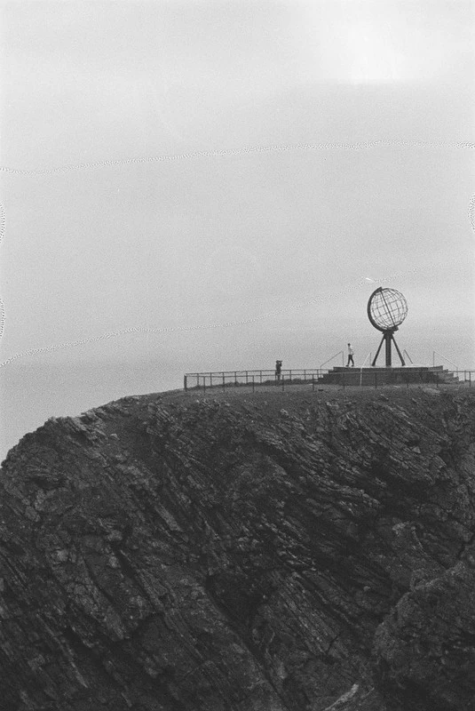 Schwarz-Weiß-Fotografie des Nordkaps: Zwei Personen stehen in der Nähe des markanten Globus-Denkmals auf einer Klippe, das von einem Geländer umgeben ist. Im Hintergrund erstreckt sich eine weite, offene Landschaft mit Blick auf das Meer.