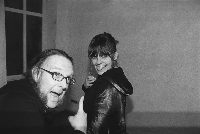 Schwarz-Weiß-Fotografie von zwei Personen in einem Innenraum: Eine Person mit Brille und Bart hält eine Zigarette und lächelt, die andere Person mit dunklen Haaren und einer Jacke steht direkt neben ihr und lächelt ebenfalls.