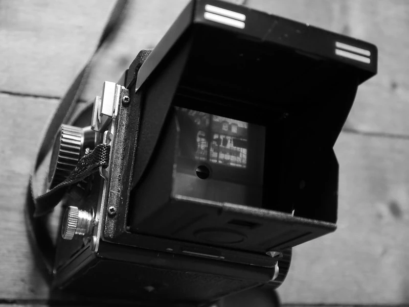 Schwarz-Weiß-Nahaufnahme einer alten Sofortbildkamera: Die Kamera ist seitlich fotografiert, der Sucher ist geöffnet und zeigt eine leere oder unscharfe Bildfläche. Die Mechanik und das Gehäuse der Kamera sind deutlich zu erkennen.