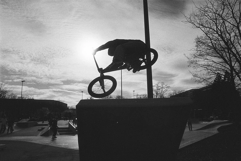 Silhouette eines BMX-Fahrers in der Luft während eines Sprungs im Skatepark bei Sonnenuntergang, im Hintergrund sind weitere Skater und Rampen zu erkennen.