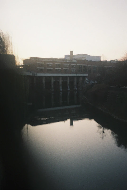 Fotografie eines ruhigen Gewässers in städtischer Umgebung: Im Vordergrund spiegelt sich ein Gebäude im Wasser, während im Hintergrund weitere Gebäude und Bäume bei Sonnenuntergang oder Sonnenaufgang zu sehen sind. Die Stimmung wirkt friedlich und leicht melancholisch.