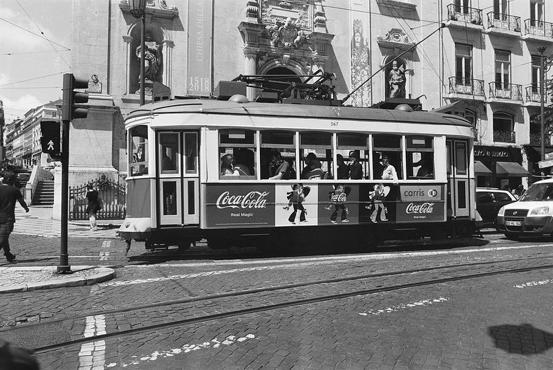Schwarz-Weiß-Fotografie einer historischen Straßenbahn in Lissabon mit Coca-Cola-Werbung, die an einer Ampel an einer belebten Straße mit portugiesischer Architektur im Hintergrund hält.