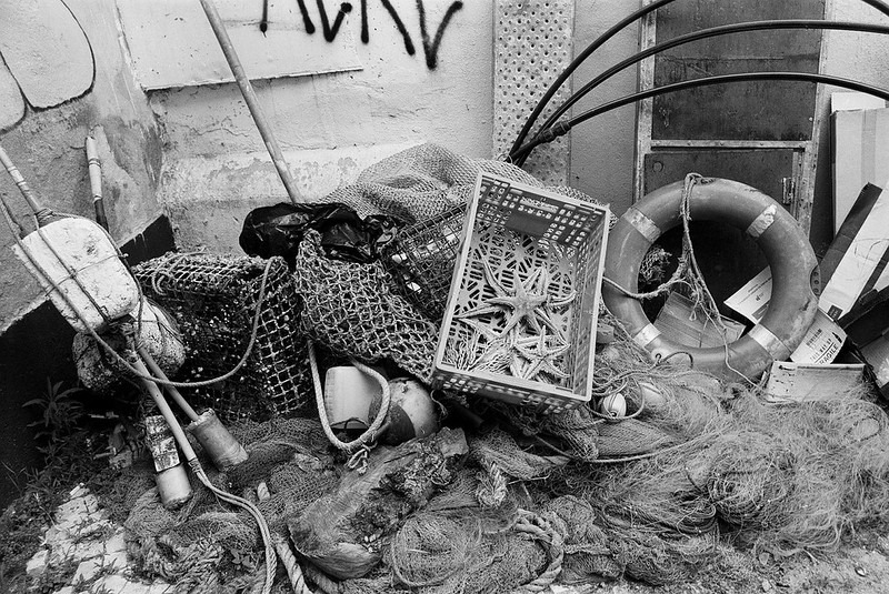 Schwarz-Weiß-Aufnahme von verstreuten Gegenständen wie Fischernetzen, einem alten Rahmen, einem Rettungsring und weiteren Fundstücken in einer Ecke, möglicherweise in einem verlassenen oder heruntergekommenen Bereich.