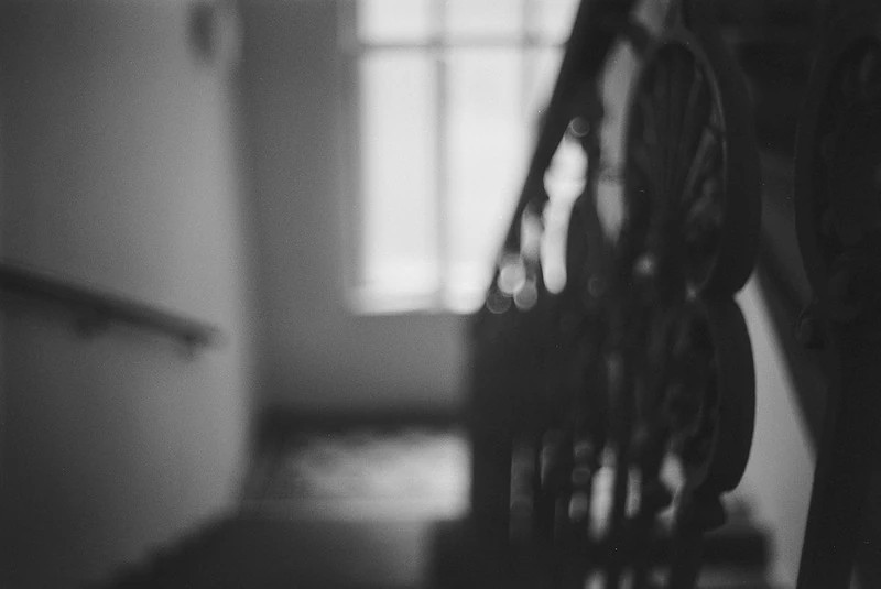 Schwarz-Weiß-Fotografie eines unscharfen Innenraums: Im Vordergrund ist ein Treppengeländer zu erkennen, im Hintergrund ein Fenster, das Licht hereinlässt. Die Szene wirkt verschwommen und abstrakt.