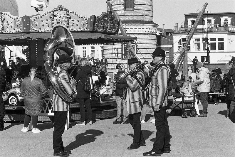 Drei Musiker in Uniformen spielen Blechblasinstrumente auf einem belebten Platz, im Hintergrund ein Karussell und historische Gebäude, fotografiert in Schwarz-Weiß.
