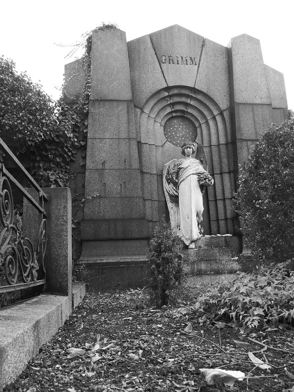 Schwarz-Weiß-Fotografie eines historischen Grabmals auf einem Friedhof: Das Grabmal ist mit dem Namen 'GRIMM' beschriftet und zeigt eine Skulptur einer stehenden Figur. Umgeben ist es von bewachsenen Sträuchern und einem schmiedeeisernen Zaun, was eine würdevoll-ruhige Atmosphäre vermittelt.