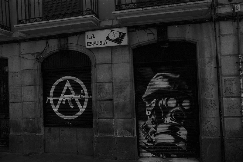Schwarz-Weiß-Fotografie einer Hausfassade mit einem Anarchie-Symbol (A im Kreis) und einem Graffiti-Porträt einer Person mit Gasmaske unter dem Schild 'La Espuela'.