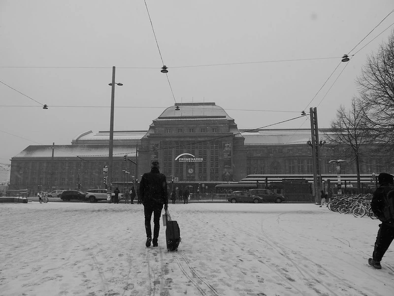 Schwarz-Weiß-Fotografie eines schneebedeckten Platzes vor einem großen, historischen Bahnhof. Zwei Personen gehen mit Gepäck durch den Schnee, während Oberleitungen und Laternenmasten im Bild zu sehen sind. Die Atmosphäre wirkt kalt und still.