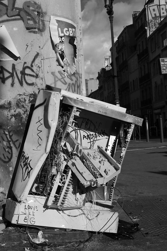 Schwarz-Weiß-Fotografie eines beschädigten Stromkastens auf einem Gehweg in einer städtischen Umgebung. Der Kasten ist aufgebrochen, mit Graffiti bedeckt und liegt zwischen Trümmern. Im Hintergrund sind Gebäude und Straßenlaternen zu sehen.