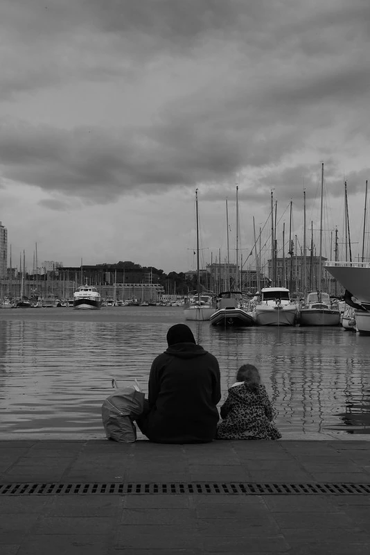 Schwarz-Weiß-Fotografie von zwei Personen, die am Ufer eines Hafens sitzen: Eine erwachsene Person und ein Kind blicken auf das Wasser, im Hintergrund liegen zahlreiche Boote und Segelmasten bei bewölktem Himmel.