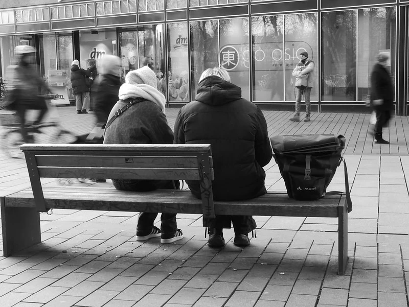Schwarz-Weiß-Fotografie einer städtischen Szene: Zwei Personen sitzen auf einer öffentlichen Bank mit dem Rücken zur Kamera. Eine der Personen hat eine große Tasche neben sich stehen. Im Hintergrund sind Passanten und ein Geschäft mit Schaufenstern zu sehen, was eine lebendige Straßenatmosphäre vermittelt.