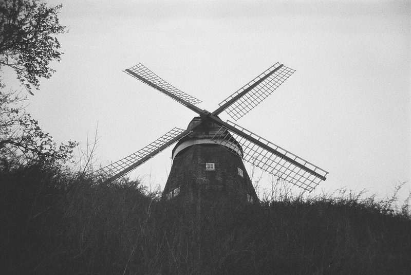 Traditionelle Windmühle mit vier Flügeln, stehend auf einem Hügel und umgeben von hohem Gras, fotografiert in Schwarz-Weiß.