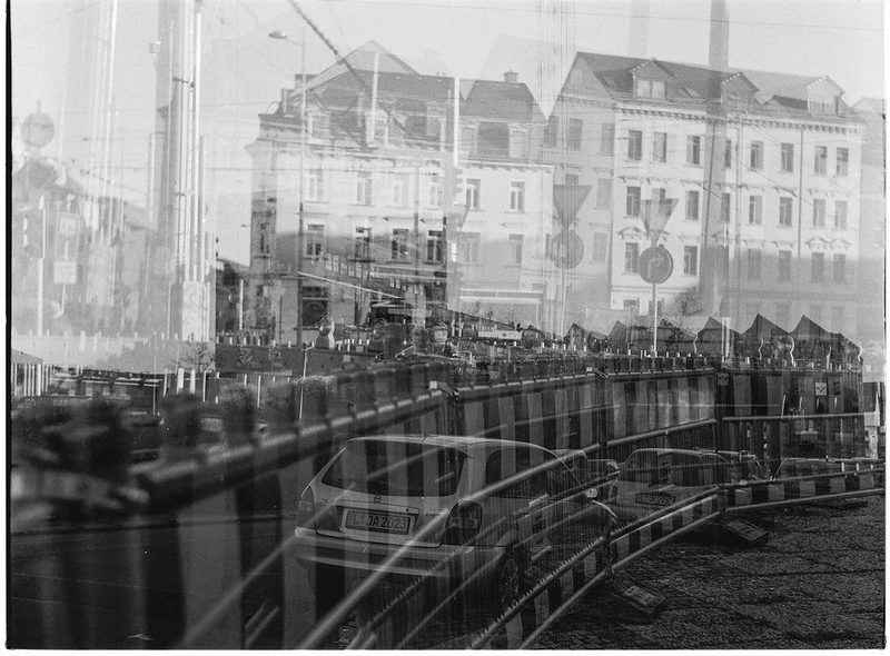 Schwarz-Weiß-Doppeltebelichtung einer städtischen Szene: Im Vordergrund sind Straßenlaternen und ein Geländer zu sehen, während sich im Hintergrund historische Gebäude und eine belebte Straße mit Menschen und Fahrzeugen überlagern. Die Aufnahme wirkt künstlerisch und surreal.