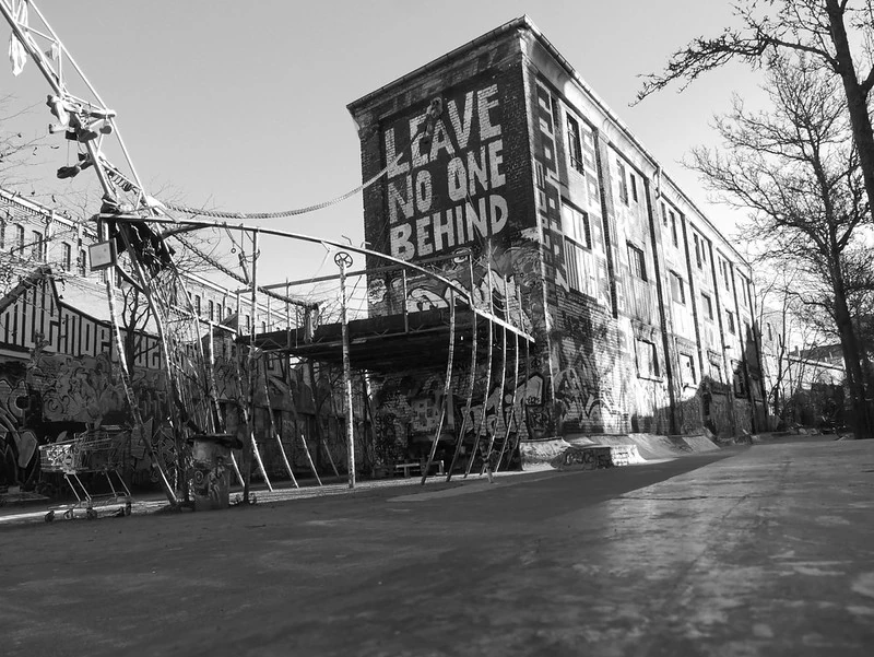 Schwarz-Weiß-Fotografie einer alten Fabrikhalle mit der Aufschrift 'LEAVE NO ONE BEHIND' an der Fassade. Im Vordergrund befindet sich eine künstlerisch gestaltete Plattform in Form eines Schiffsbugs. Die Szene wirkt verlassen und künstlerisch."