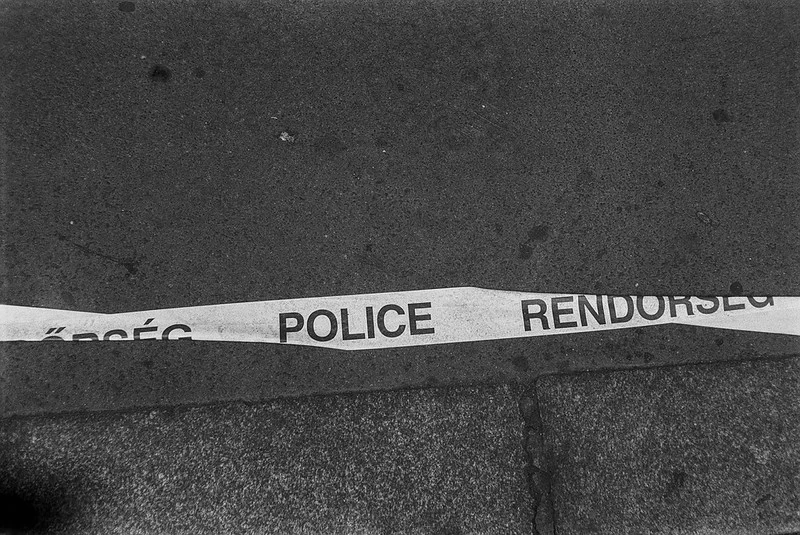 Schwarz-Weiß-Fotografie: Ein Polizeiabsperrband liegt auf dem Boden; darauf ist das Wort 'POLICE' lesbar, das nächste Wort ist nur teilweise erkennbar.