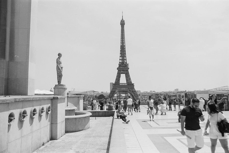 Schwarz-Weiß-Fotografie des Eiffelturms in Paris, aufgenommen von einer erhöhten Position mit Blick auf die Menschenmenge und eine Statue im Vordergrund.
