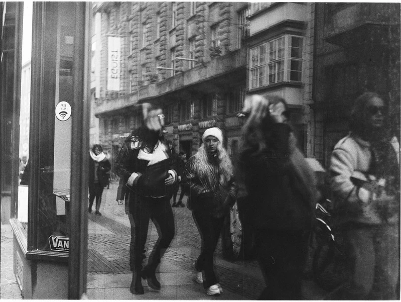 Schwarz-Weiß-Fotografie einer Spiegelung in einer Schaufensterscheibe: Die Reflexion zeigt Passanten auf einer belebten Stadtstraße, die an Gebäuden und Geschäften vorbeigehen. Die Szene wirkt lebhaft und dynamisch.