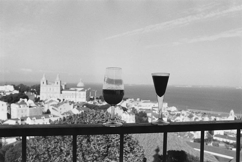 Schwarz-Weiß-Fotografie mit Blick auf den Lissabon-Hafen: Im Vordergrund zwei kleine Gläser mit Portwein, im Hintergrund mehrere Boote im Wasser und die Skyline der Stadt mit historischen Gebäuden.